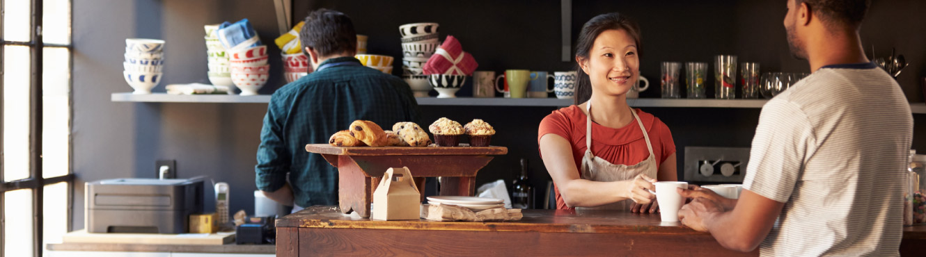 Cafe owner serves a cup of coffee to a man at the counter with an employee working behind her 