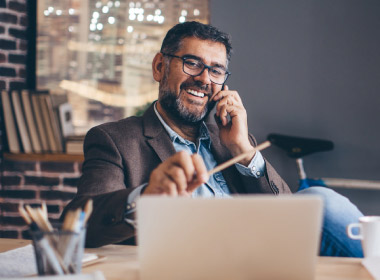 Casual business man sits at his desk, talking on the phone and looking at his computer