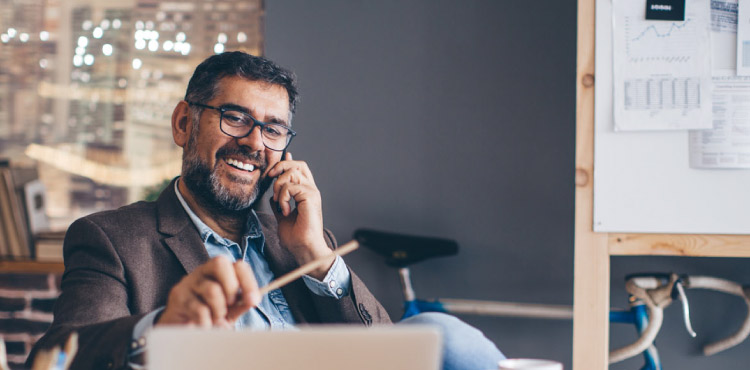 Casual business man sits at his desk, talking on the phone and looking at his computer