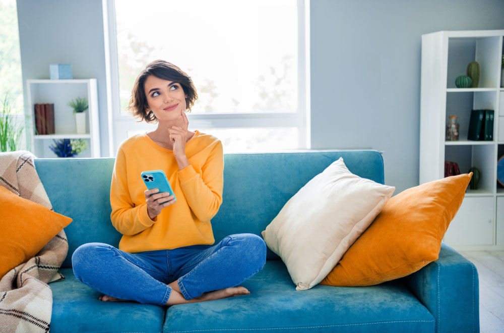 Person sitting on a couch using their phone for digital banking and thinking about their finances.