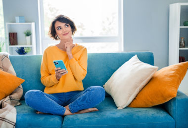 Person sitting on a couch using their phone for digital banking and thinking about their finances.
