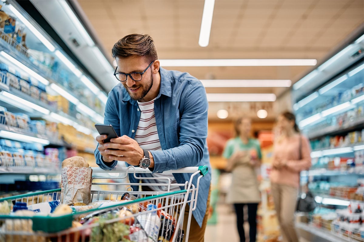 Happy man going through checklist on smart phone while shopping in supermarket.